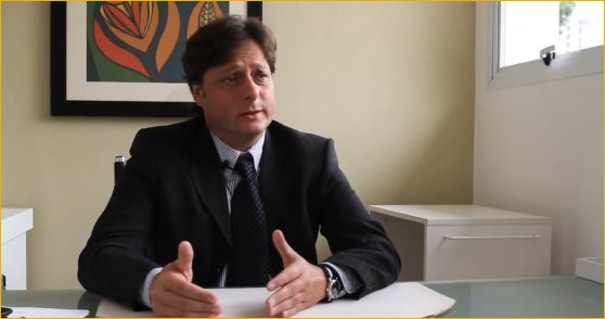 Imagem do Doutor Mauad de terno preto explicando alguma coisa em sua mesa de atendimento